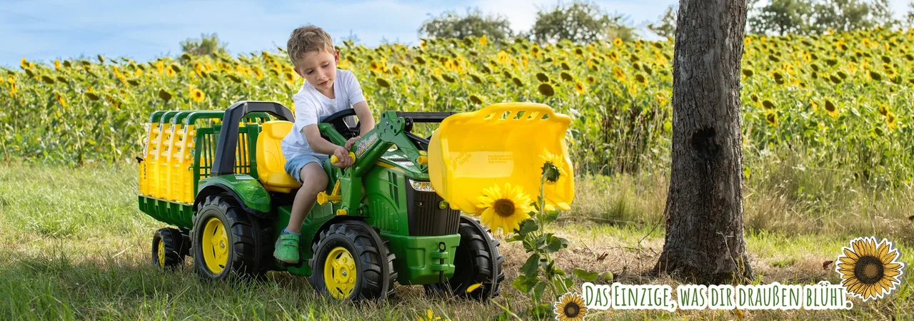 Kind auf einem rolly toys Trettraktor vor einem Sonnenblumenfeld